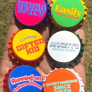 May include: Six circular bottle caps featuring colorful designs and text. The caps display the phrases: Neuro Spicy, Easily Distracted, Former Gifted Kid, Anxiety's Employee of the Month, Running on Caffeine & Chaos, and Overthinking Since Birth.
