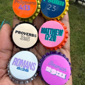 May include: Six circular bottle caps featuring religious text. The caps are orange, yellow, black, orange, silver, and purple. The text includes Philippians 4:13, Psalm 23, Proverbs 3:5, Matthew 17:20, Romans 8:28, and Jeremiah 29:11 in various colors.