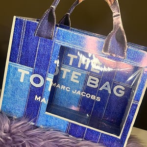May include: A blue denim-style tote bag with the text "THE TOTE BAG" and "MARC JACOBS" in white. The bag has a clear window and brown and blue handles. The bag is sitting on a purple furry surface.
