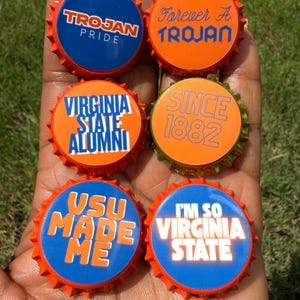 May include: Six circular bottle caps featuring orange and blue designs with text including Trojan Pride, Forever A Trojan, Virginia State Alumni, Since 1882, VSU Made Me, and I'm So Virginia State, arranged in two columns on a palm.