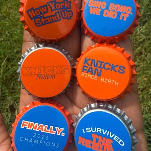 May include: Six circular metal bottle caps featuring orange and blue designs with text including New York Stand Up, Bing Bong We Did It, Knicks Nation, Knicks Fan Since Birth, Finally 2026 Champions, and I Survived The Rebuild.