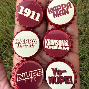 May include: Six circular bottle caps featuring a crimson and cream color scheme. The caps display text including 1911, KAPPA MAN, KAPPA Made Me, KRIMSON & KREAM, NUPE LIFE, and Yo-NUPE! The designs are printed on a glossy surface with serrated edges.