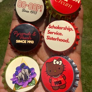 May include: Six decorative bottle caps featuring various designs including text that reads 00-OOP! Since 1913, Crimson & Cream, Pyramids & Pearls SINCE 1913, Scholarship. Service. Sisterhood., an elephant on a purple flower, and a cartoon cat with Diva Since 1913.