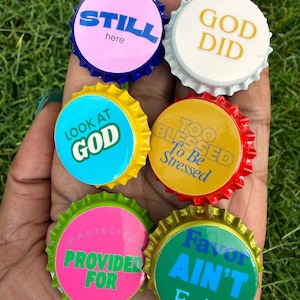 May include: Six circular metal bottle caps with serrated edges, each featuring unique text. The caps are colored pink, white, blue, red, neon pink, and green, with printed phrases including STILL here, GOD DID, LOOK AT GOD, TOO BLESSED To Be Stressed, PROTECTED PROVIDED FOR, and Favor AIN'T Fair.