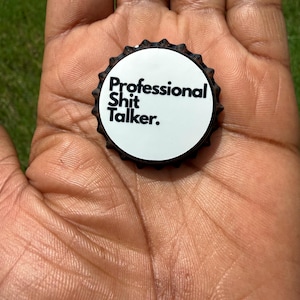 May include: A black metal bottle cap with a white circular center featuring the black text Professional Shit Talker. The cap has a serrated edge and a standard crimped design, presenting a bold and direct message in a compact, circular format.
