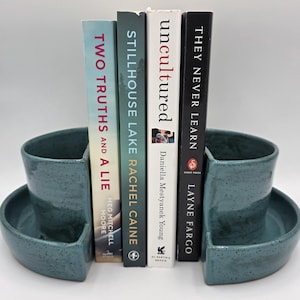 May include: A pair of teal ceramic bookends with a speckled finish, holding several books. The books have titles like "Two Truths and a Lie" and "Stillhouse Lake." The bookends have a curved design with a flat base.