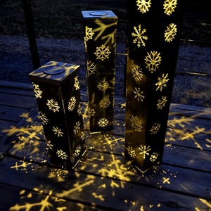 May include: Three black, rectangular outdoor lanterns with snowflake cutouts. The lanterns are illuminated from within, casting snowflake-shaped shadows on the wooden deck. The lanterns are of varying heights and have a square top.
