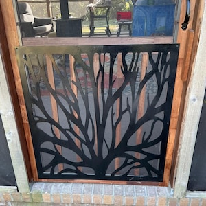 May include: A black metal screen door with a tree branch design. The door is framed in wood and has a handle on the right side. The screen door is installed on a brick surface.