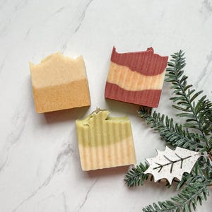 May include: Three bars of handmade soap, one is green and white, one is red and white, and one is yellow and white. The soaps are on a white marble surface.
