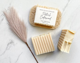 May include: Two bars of goat milk soap with a label that reads "Hope Filled With Joy Patchouli Cedarwood Goat Milk Soap Approx. 4 oz". The soap is on a white marble surface with a natural loofah sponge and a light brown feather duster.
