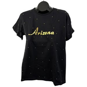 Puede incluir: Camiseta negra con la palabra "Arizona" en letras doradas. La camiseta está adornada con pequeños tachuelas doradas dispersas por la parte delantera. Tiene mangas cortas y cuello redondo.