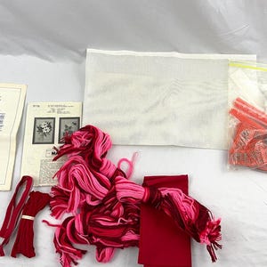 Könnte beinhalten: Ein Super Craft Kit mit Anleitungen für verschiedene Bastelarbeiten, darunter Sticken und Stricken. Das Set enthält rote und rosa Wolle, rote Bänder, ein weißes Stoffstück und eine Tüte mit roten Kunststoffteilen.