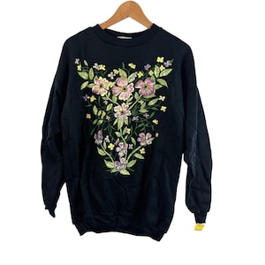 Puede incluir: Sudadera negra con un diseño floral bordado. El diseño presenta flores rosas, amarillas y moradas con hojas y tallos verdes. La sudadera está colgada en una percha de madera.
