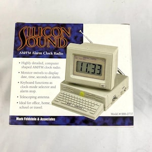 May include: A beige AM/FM alarm clock radio shaped like a computer, with a digital display showing 11:33. The clock has a keyboard-like base and a telescoping antenna. The box reads "SILICON SOUND" and "AM/FM Alarm Clock Radio".