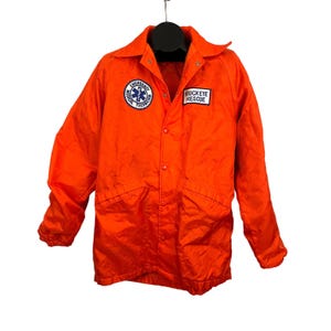 May include: Orange jacket with a snap closure and two pockets. The jacket has a patch on the left side that says "Emergency Medical Technician" and a patch on the right side that says "Buckeye Rescue".