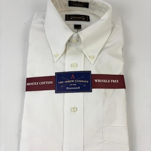 Może przedstawiać: Złożona biała koszula z kołnierzykiem zapinanym na guziki i kieszenią na piersi. Koszula jest zapakowana z bordową opaską z napisem "Mostly Cotton" i "Wrinkle Free". Niebieska etykieta wyświetla "The Arrow Company" i "Pinpoint".