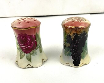 Puede incluir: Un par de saleros y pimenteros vintage con diseños florales. El salero de la izquierda tiene un diseño de rosa rosa, mientras que el salero de la derecha tiene un diseño de uva negra.