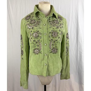 Pode incluir: Uma camisa de camurça verde claro com intrincados bordados e contas prateadas e transparentes. Os enfeites estão concentrados ao redor da gola, da frente e das mangas. A camisa é exibida em um manequim.