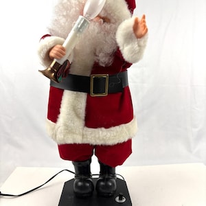 Puede incluir: Una figura de Papá Noel roja y blanca sosteniendo una luz blanca con forma de vela. La figura tiene barba blanca y lleva cinturón y botas negras. La figura está sobre una base negra con un interruptor.