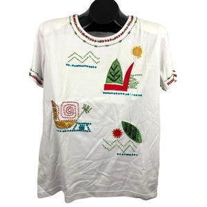 May include: White t-shirt with colorful embroidered designs. The shirt features a boat, sun, and tree motifs, along with geometric patterns. The neckline and sleeves are trimmed with red and green accents. A unique, artistic garment.