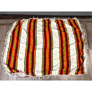May include: A white blanket with vertical stripes in red, orange, yellow, and green. The blanket has a fringe border. The blanket is lying on a wooden floor.
