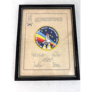 May include: A framed display featuring a circular Space Shuttle Atlantis mission patch with a rainbow trail and stars, surrounded by the names Mullane, Ross, Shepherd, Gibson, and Gardner. Below are five astronaut signatures on a beige background.