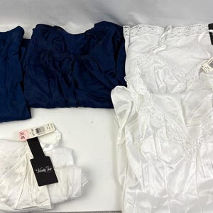 Könnte beinhalten: Eine Auswahl an Dessous, darunter zwei marineblaue Oberteile und mehrere weiße Satin-Unterkleider mit Spitzenbesatz. Jedes Teil hat ein Etikett. Die Artikel sind auf einer weißen Oberfläche angeordnet.