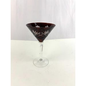 May include: A burgundy cocktail glass with a clear stem and base. The glass features a triangular bowl with etched floral patterns. The stem is clear, and the base is round. The glass is set against a white background.
