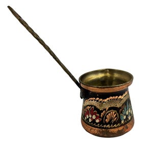 May include: A small, copper-colored coffee pot with a long, twisted handle. The pot features a black base with floral designs in red, gold, and blue. The interior of the pot is gold-colored.