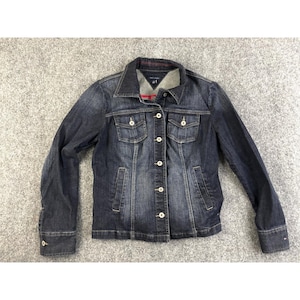 Puede incluir: Chaqueta vaquera azul oscuro con cierre de botones, dos bolsillos en el pecho y dos bolsillos laterales. La chaqueta tiene un cuello clásico y mangas largas con puños abotonados. El logo de Tommy Hilfiger es visible en el puño.