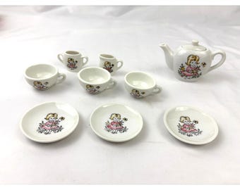 May include: A miniature white ceramic tea set, including a teapot, cups, saucers, and cream jugs. Each item is adorned with a cartoon girl in a pink dress. Ideal for children's play or as a decorative item.