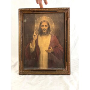 May include: Framed religious artwork featuring a portrait of Jesus Christ. The image shows Jesus with a halo, wearing a red robe and a cream-colored tunic. The artwork is in a wooden frame.