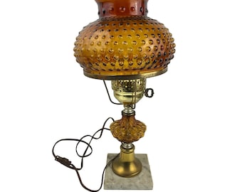 Może przedstawiać: Lampa stołowa w stylu vintage wykonana z bursztynowego szkła z fakturą w postaci bąbelków. Lampa ma złotą metalową podstawę i białą marmurową podstawę.