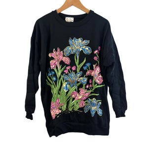 Puede incluir: Sudadera negra de manga larga con un diseño floral de iris en tonos azules, rosas y verdes. Los iris están bordados en la parte delantera de la sudadera. La sudadera tiene cuello redondo y mangas largas.
