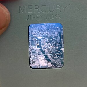 Könnte beinhalten: Ein Vintage-Dia mit grauem Hintergrund und dem in Weiß gedruckten Text "MERCURY JIFFY-MASK". Das Dia enthält ein Foto einer Luftaufnahme einer Stadt mit Gebäuden und Straßen.