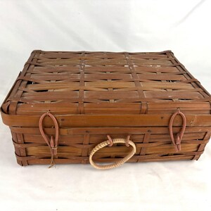May include: A rectangular, woven brown basket with a lid. The basket has a woven handle and two leather-like straps with loops. The basket is made of woven wood strips and is likely used for storage or carrying items.