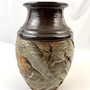 May include: A handcrafted ceramic vase with a dark brown rim and a textured, light brown and gray body. The vase has a wide base and a narrower neck, showcasing a unique, organic design. The vase is suitable for home decor.