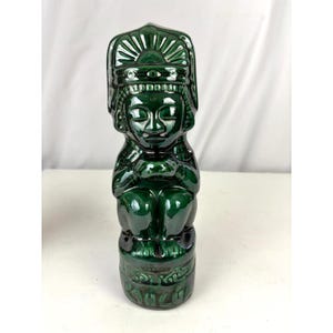 May include: A dark green ceramic figurine of a seated figure with a detailed headdress. The figurine has a glossy finish and is approximately 20 cm tall. The figure is seated on a pedestal with carved details.
