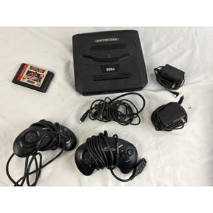 May include: A black Sega Genesis video game console with two controllers, power adapters, and a game cartridge. The console has the word "Genesis" printed on the top and "Sega" on the front. The game cartridge has the word "Sega" printed on it.