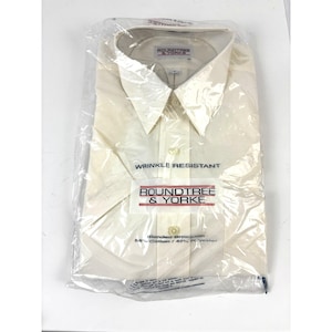 Pode incluir: Uma camisa social creme de manga comprida da Roundtree & Yorke, embalada em um saco plástico transparente. A camisa é rotulada como "Wrinkle Resistant" e feita de tecido misto. A embalagem inclui o logotipo da marca e detalhes da composição do tecido.