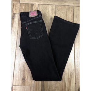 May include: Black Levi's jeans with a flared leg. The jeans have a classic five-pocket design with white stitching and a red Levi's tag on the back pocket. The Levi's label is pink. The jeans are laid flat on a wooden surface.