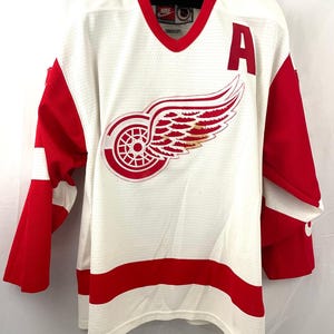 Könnte beinhalten: Ein weißes Hockeytrikot mit roten und weißen Streifen. Das Trikot hat ein rotes Flügelrad-Logo auf der Vorderseite und ein rotes "A" auf der rechten Schulter. Das Trikot ist von Nike.