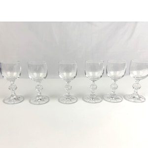 Könnte beinhalten: Sechs klare Likörgläser. Jedes Glas hat eine runde Schale, einen dekorativen Stiel mit Facettendesign und einen kreisförmigen Fuß. Die Gläser sind in einer Reihe vor weißem Hintergrund angeordnet.