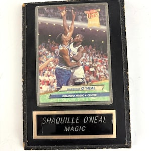 May include: A framed basketball card featuring Shaquille O'Neal in action for the Orlando Magic. The card is set against a black background with a gold-coloured nameplate that reads "Shaquille O'Neal Magic."