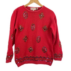 Può includere: Un maglione rosso con perline decorative e abbellimenti di paillettes. Il maglione ha un girocollo e maniche lunghe. Il design include varie forme di motivi con perline e paillettes. Il marchio "Carlucci" è visibile.