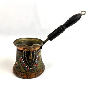 May include: A vintage copper coffee pot with a black wooden handle. The pot features ornate detailing with colorful accents of red, blue, and yellow. The pot is designed with a long handle and a small spout.
