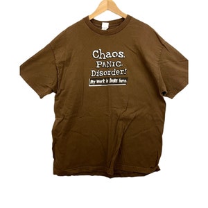 Könnte beinhalten: Braunes T-Shirt mit weißem Text, der lautet: "Chaos. Panik. Störung! Meine Arbeit ist HIER GESCHEHEN."