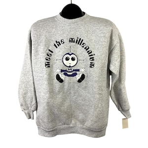 Puede incluir: Una sudadera gris con la frase "meet the millennium" arqueada sobre un robot de dibujos animados. El robot es azul, blanco y negro con ojos grandes y brazos extendidos. Una etiqueta cuelga de la manga.