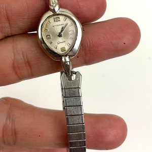 May include: Vintage Wittnauer Genève watch with a silver-toned oval face and a silver-toned expanding band. The watch face displays the number 12, and the word "Wittnauer" is printed above "Genève". The watch is held by a hand.