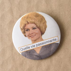 Peut inclure: Un bouton rond blanc avec une image vintage d'une femme aux cheveux blonds. Le bouton porte le texte "Quelle vie passionnante." dans un encadré rectangulaire. Le fond est brun clair.